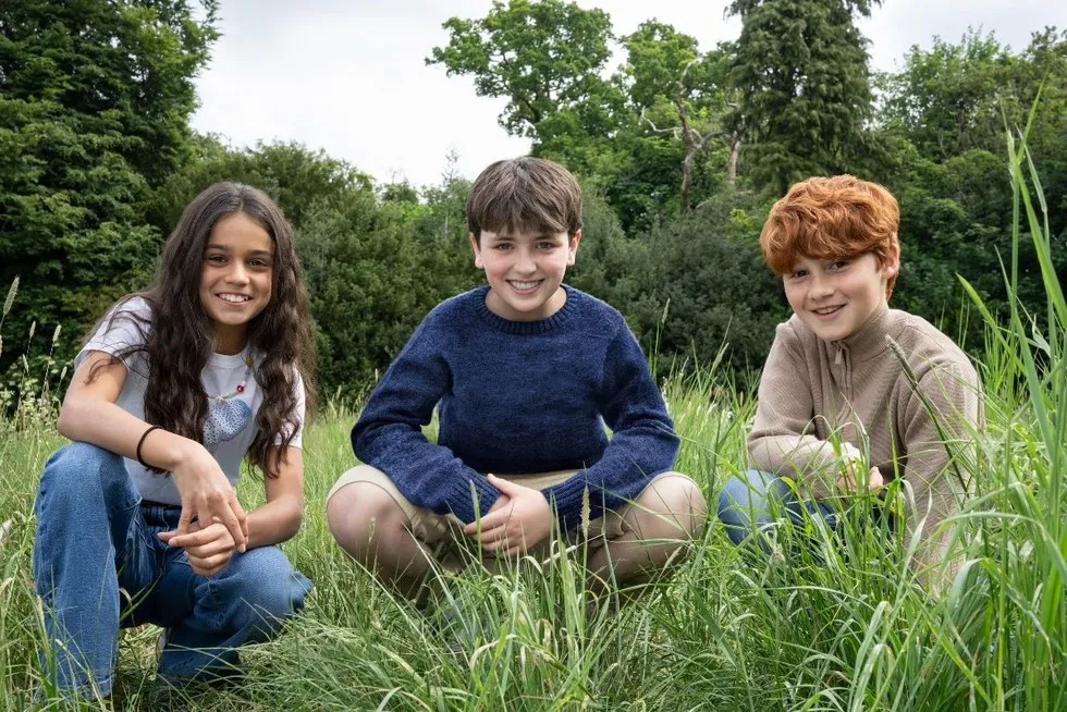 Dominic McLaughlin, Arabella Stanton and Alastair Stout crouch in some grass in a field outdoors, smiling at the camera.
